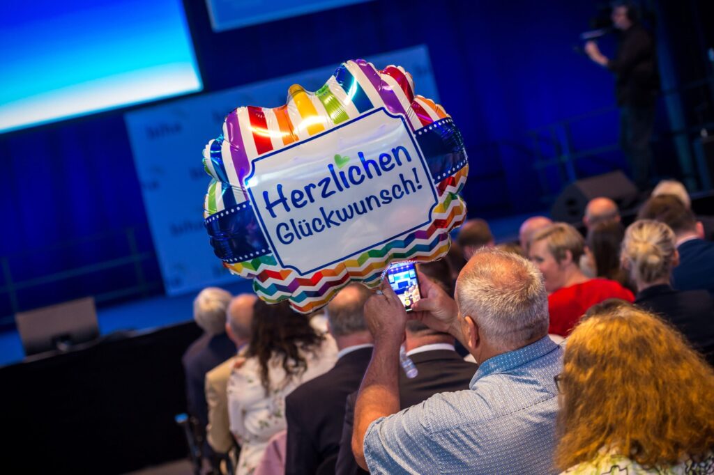 m Vordergrund hält eine Person einen bunten Folienballon mit der Aufschrift 'Herzlichen Glückwunsch!' in die Höhe und fotografiert ihn mit einem Smartphone. Der Ballon ist mit Regenbogenfarben und Zickzackmustern verziert. Im Hintergrund sitzen mehrere Menschen in Reihen, die Bühne ist blau ausgeleuchtet.