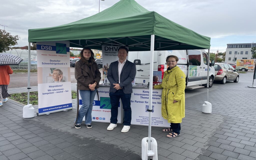 Drei Personen stehen unter einem grünen Pavillon mit Informationsständen des Deutschen Schwerhörigenbunds und Deutschen Hörverbands auf einem gepflasterten Platz.