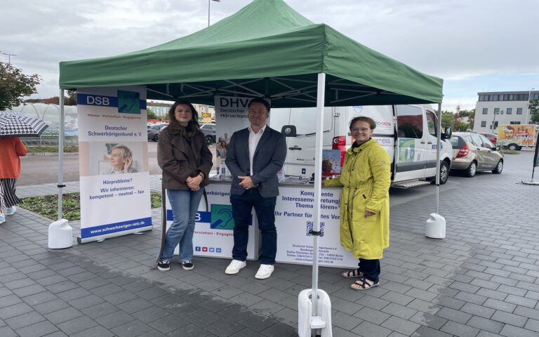 Drei Personen stehen unter einem grünen Pavillon mit Informationsständen des Deutschen Schwerhörigenbunds und Deutschen Hörverbands auf einem gepflasterten Platz.