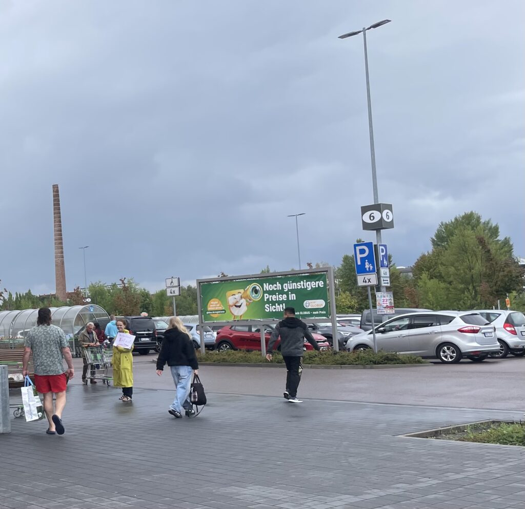 Menschen mit Einkaufstaschen und Einkaufswagen auf einem Parkplatz unter bewölktem Himmel mit Werbetafel im Hintergrund.