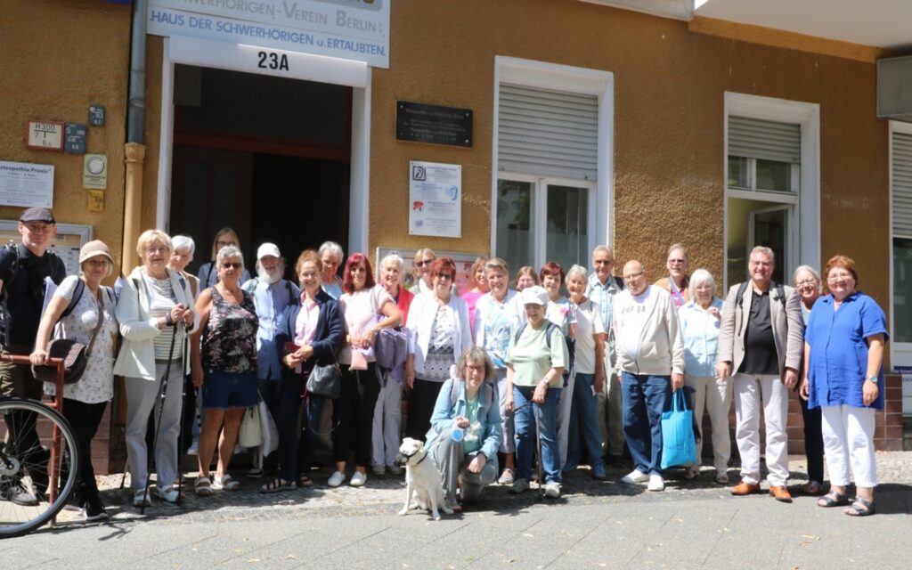 Gruppe von Menschen unterschiedlichen Alters steht vor einem Gebäude mit gelber Fassade und mehreren Fenstern