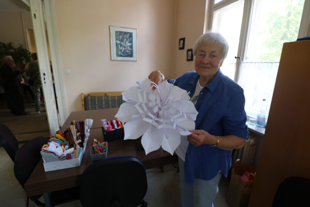 Eine ältere Frau steht in einem hellen Raum und hält eine große, weiße Papierblume in der Hand. Auf dem Tisch neben ihr liegen Bastelmaterialien wie bunte Stifte, Scheren und Papierrollen. Im Hintergrund sind ein Fenster, ein Schrank und ein gerahmtes Bild an der Wand zu sehen. Die Frau trägt eine blaue Jacke und lächelt leicht.