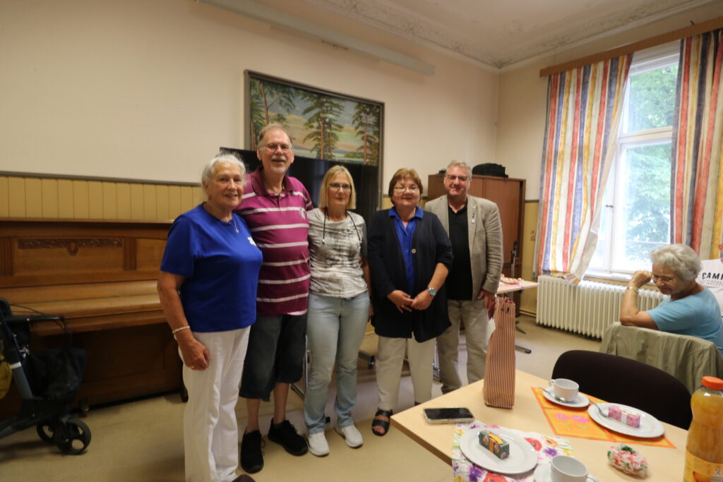 ünf Personen stehen nebeneinander in einem hellen Raum mit hohen Decken. Im Hintergrund hängt ein großes Gemälde mit Landschaftsmotiv an der Wand. Links steht ein Klavier, daneben ein Rollator. Rechts im Bild ist ein Fenster mit gestreiften Vorhängen zu sehen. Auf dem Tisch im Vordergrund stehen Kaffeetassen, Teller mit Gebäck, eine Flasche Orangensaft und eine dekorative Flasche mit Stoffüberzug. Die Personen tragen Freizeitkleidung und lächeln in die Kamera.