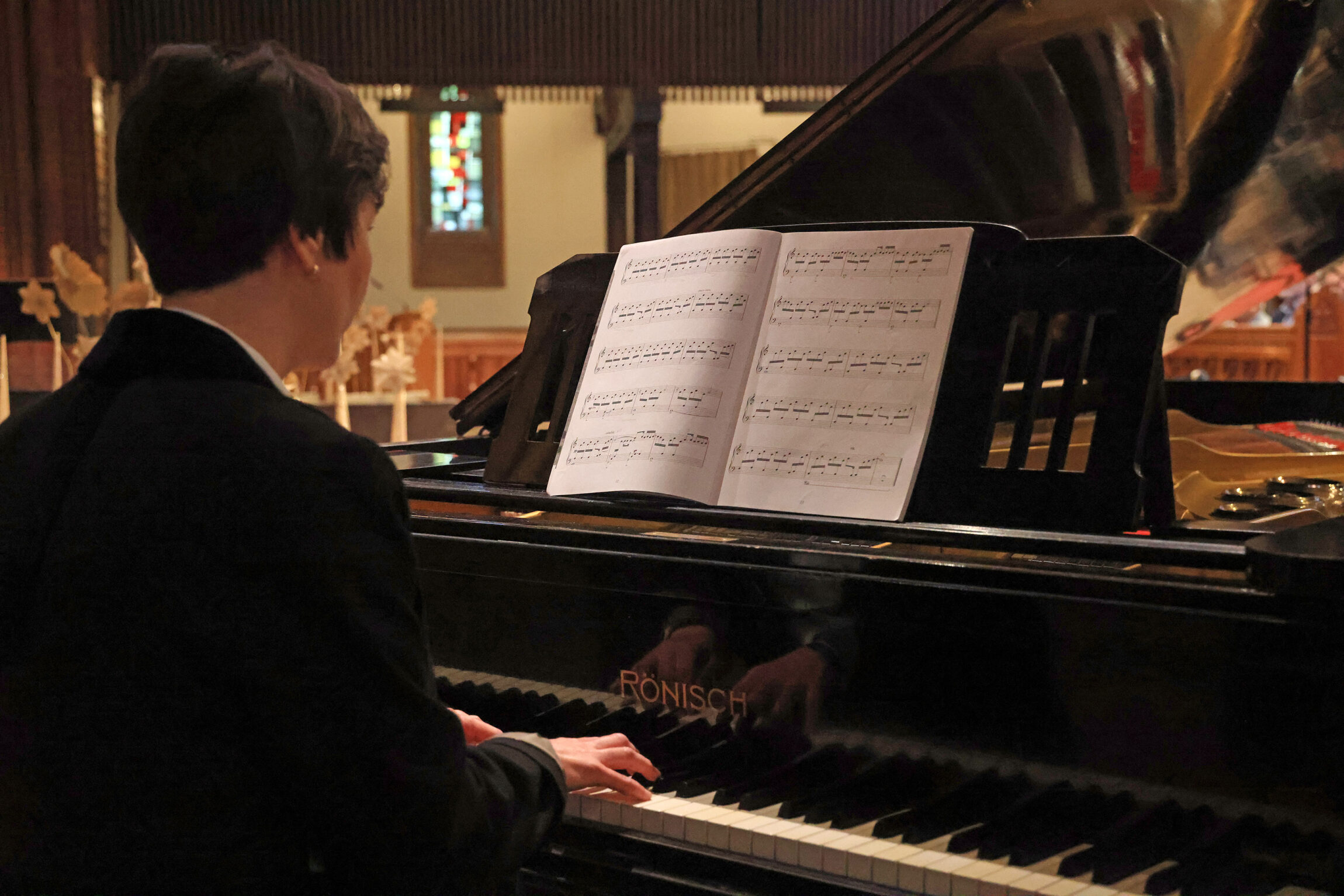 Eine Person sitzt an einem Flügel und spielt Klavier. Auf dem Instrument liegt ein aufgeschlagenes Notenblatt, dessen Notenlinien und Symbole deutlich erkennbar sind. Im Hintergrund sind Holzelemente und ein buntes Fenster mit Glasmalerei zu sehen. Die Szene wirkt ruhig und konzentriert.