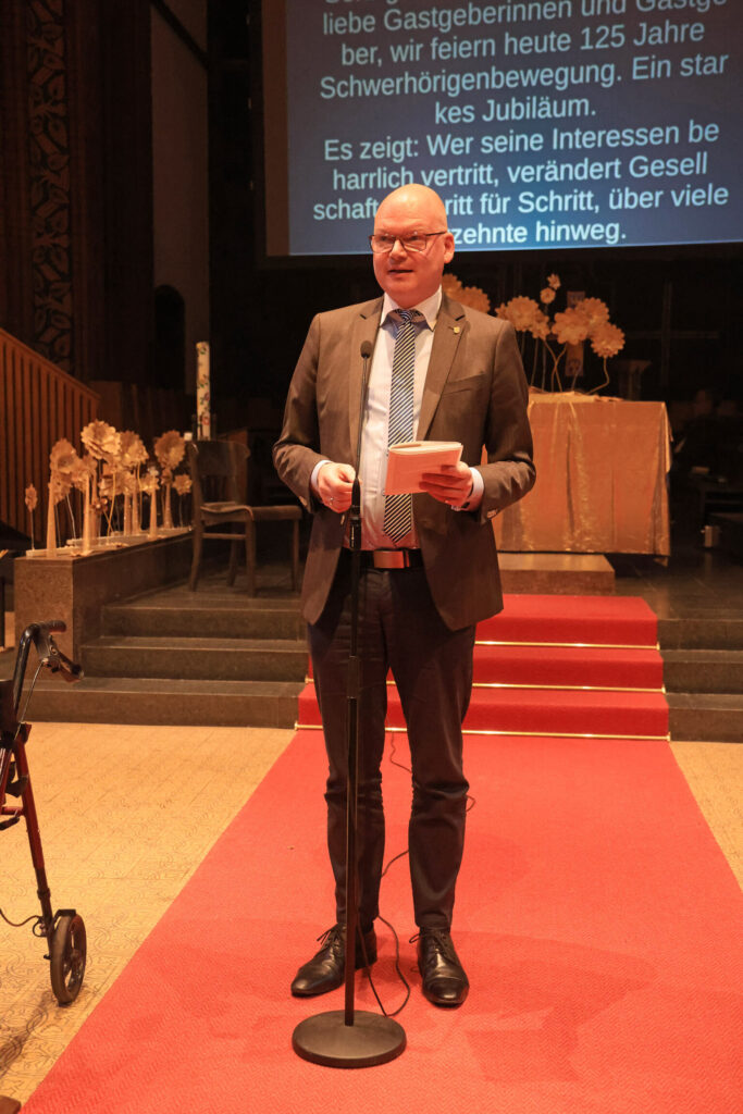 Ein Mann mit Brille und Anzug steht auf einem roten Teppich und spricht in ein Mikrofon. Er hält ein Blatt Papier in der Hand. Im Hintergrund sind goldfarbene Dekorationen und ein Tisch mit beigem Tuch zu sehen. Links im Bild steht ein Rollator. Auf einer Leinwand hinter dem Mann ist ein Text in weißer Schrift zu erkennen.