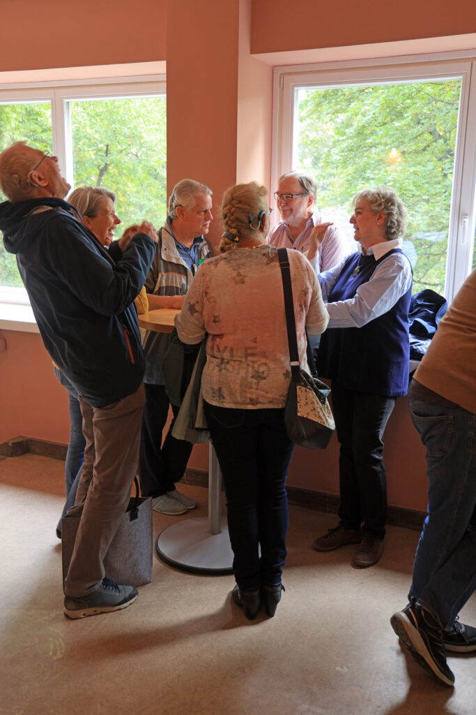 Gruppe älterer Menschen steht in einem Raum mit großen Fenstern und unterhält sich stehend um einen kleinen runden Tisch.
