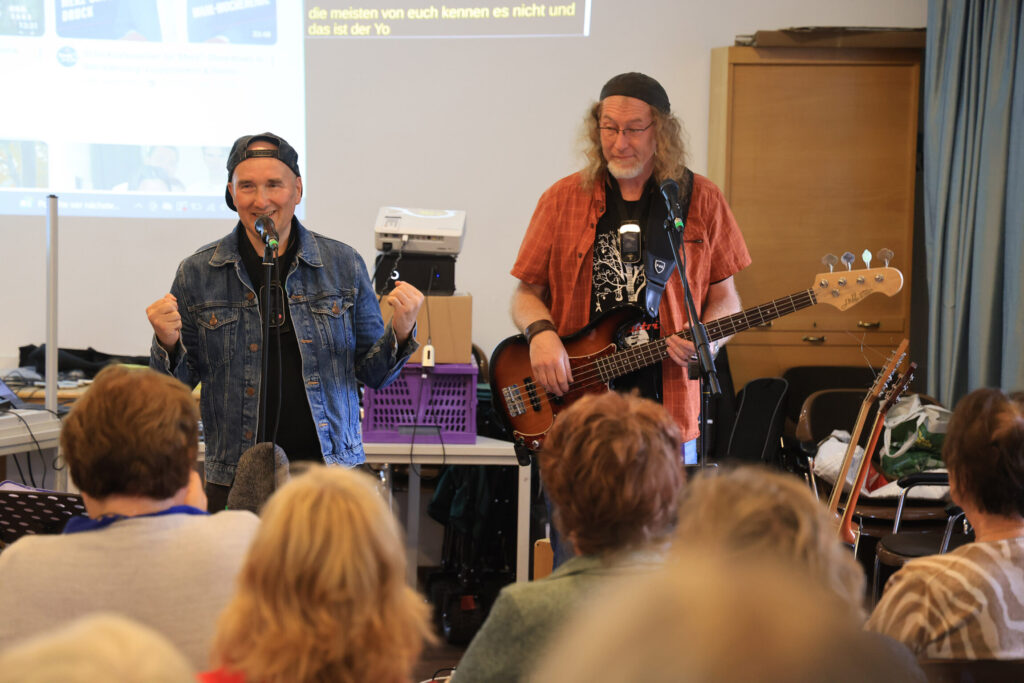 Zwei Männer stehen vor Publikum, einer spricht in ein Mikrofon, der andere spielt E-Bass.
