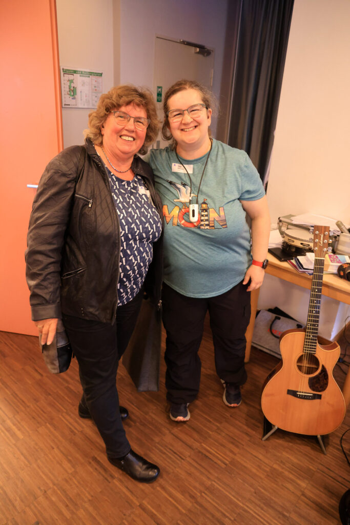 Zwei lächelnde Frauen stehen nebeneinander in einem Raum mit Holzboden, rechts eine Akustikgitarre auf einem Ständer.