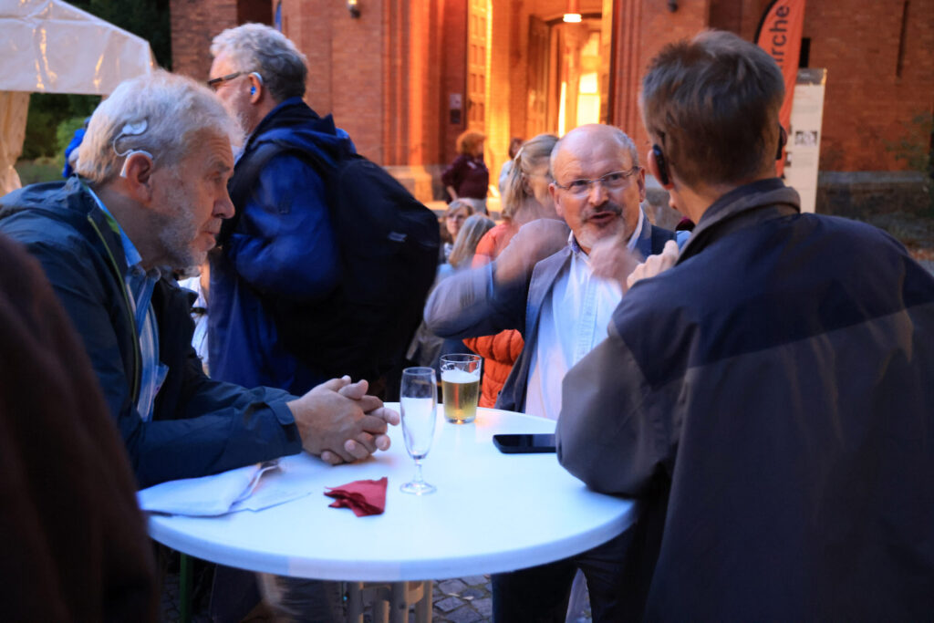 Drei Männer unterhalten sich an einem Stehtisch im Freien vor einem Backsteingebäude.