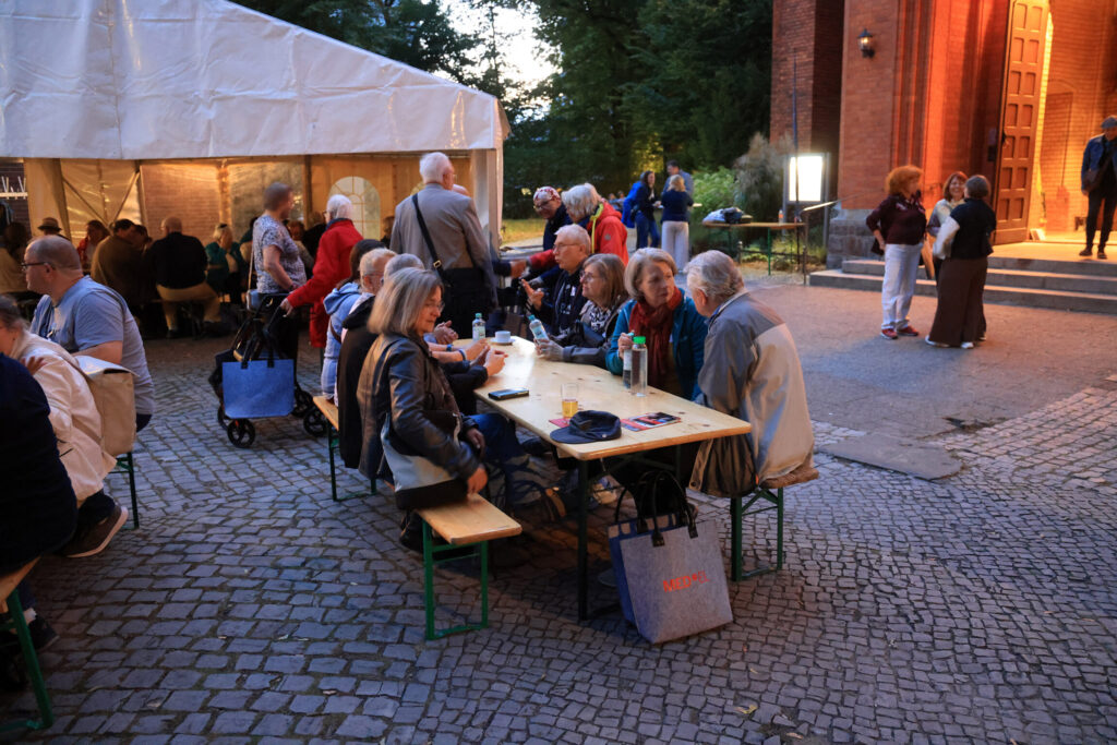 Im Vordergrund sitzen mehrere ältere Menschen an einem langen Holztisch mit Bänken auf einem gepflasterten Platz. Sie unterhalten sich und trinken aus Flaschen und Gläsern. Im Hintergrund steht ein weißes Festzelt, daneben befindet sich ein Backsteingebäude mit einer geöffneten Tür, vor der einige Personen stehen. Die Szene spielt in der Dämmerung, das Licht wirkt warm und einladend.