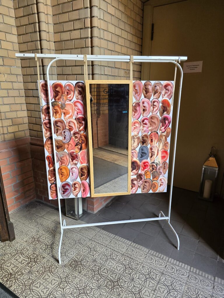 Spiegel mit weißen Metallrahmen, an dessen Seiten zahlreiche Ohrenbilder in verschiedenen Hauttönen befestigt sind, steht vor einer Ziegelwand auf einem gemusterten Boden.