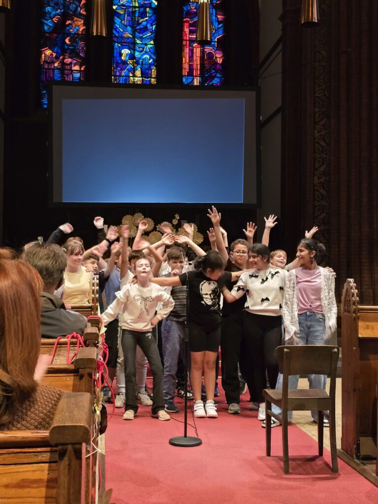 Eine Gruppe von Kindern steht auf einem roten Teppich in einer Kirche vor einem Mikrofon. Die Kinder heben die Arme und lächeln. Im Hintergrund sind bunte Glasfenster und eine große Leinwand zu sehen. Die Sitzreihen und Holzbänke sind teilweise besetzt.