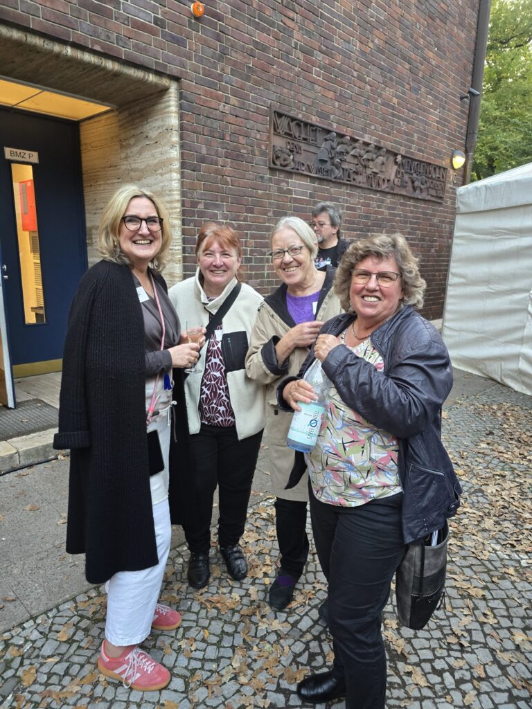 Vier Frauen stehen lachend vor einem Backsteingebäude auf einem mit Laub bedeckten Gehweg, eine hält eine Wasserflasche.