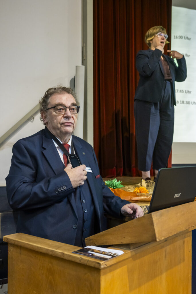 Ein Mann im dunklen Anzug steht hinter einem Holzpult und spricht, während er auf einen Laptop blickt. Im Hintergrund steht eine Frau auf einer Bühne vor einem roten Vorhang und übersetzt in Gebärdensprache. Auf der weißen Wand hinter ihr sind Uhrzeiten und Text in schwarzer Schrift zu erkennen.