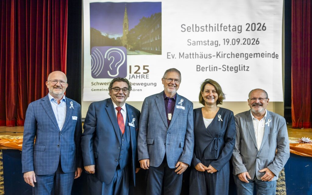 Fünf Personen stehen nebeneinander vor einer Bühne mit rotem Vorhang. Alle tragen formelle Kleidung und ein weißes Schleifenband am Revers. Im Hintergrund ist eine große Leinwand mit einer Projektion zu sehen. Links zeigt die Projektion ein Logo mit einer stilisierten Ohrmuschel und dem Text '125 Jahre Schwerhörigenbewegung'. Rechts stehen die Worte 'Selbsthilfetag 2026', das Datum 'Samstag, 19.09.2026' und der Veranstaltungsort 'Ev. Matthäus-Kirchengemeinde Berlin-Steglitz'.