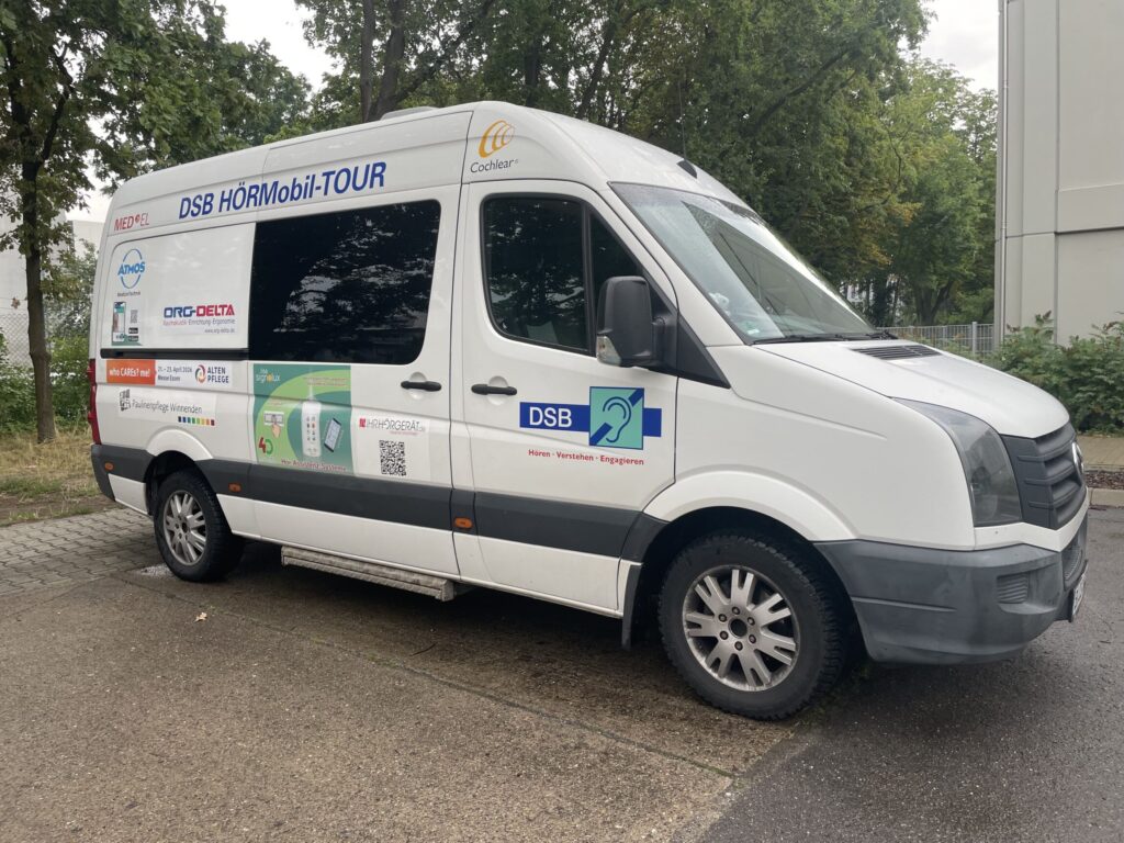 Ein weißer Transporter mit auffälligen Aufklebern und Beschriftungen steht auf einem Parkplatz. Die Seitenfläche zeigt mehrere Logos, einen QR-Code und den Schriftzug 'DSB HÖRMobil-TOUR'. Auf der Fahrerseite sind weitere Sponsorenlogos und ein großes Symbol mit einem Ohr abgebildet. Im Hintergrund sind Bäume und ein Gebäude zu sehen.