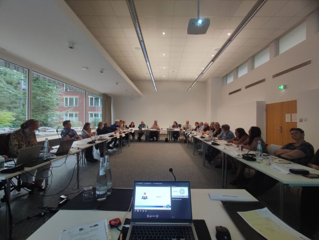 Konferenzraum mit langen weißen Tischen, die in U-Form angeordnet sind. Zahlreiche Personen sitzen an den Tischen, viele mit Laptops oder Notizblöcken. Im Vordergrund steht ein Laptop auf einem Tisch, dessen Bildschirm Präsentationsfolien zeigt. Auf dem Tisch liegen außerdem eine Wasserflasche und ein Mikrofon. Große Fenster auf der linken Seite lassen Tageslicht herein und geben den Blick auf ein Gebäude mit roten Ziegeln und grünen Bäumen frei. Die Decke ist mit eingelassenen Leuchten ausgestattet.
