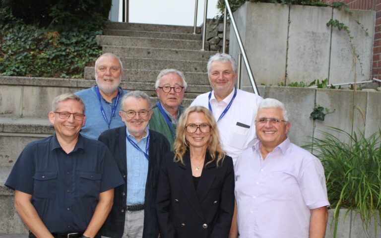 Gruppe von sieben Personen unterschiedlichen Alters vor einer Treppe und einer Betonwand, alle lächelnd und mit Namensschildern an blauen Bändern.