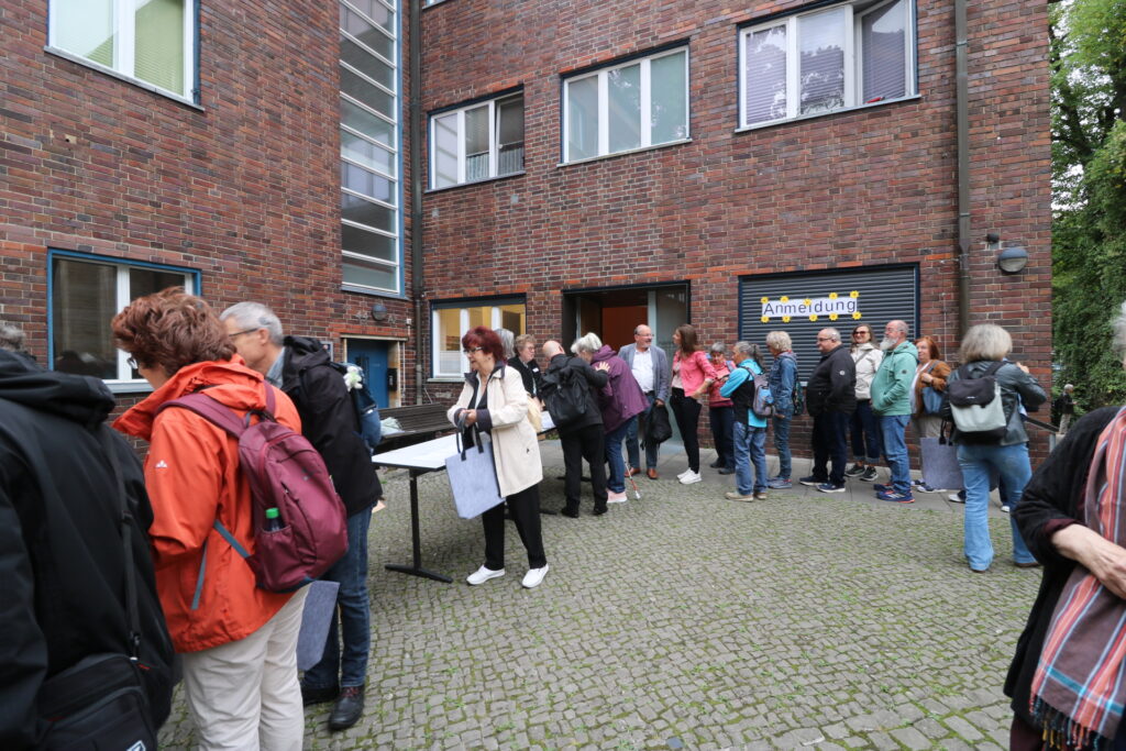 Gruppe von Menschen steht vor einem Backsteingebäude mit einem Schild 'Anmeldung'.