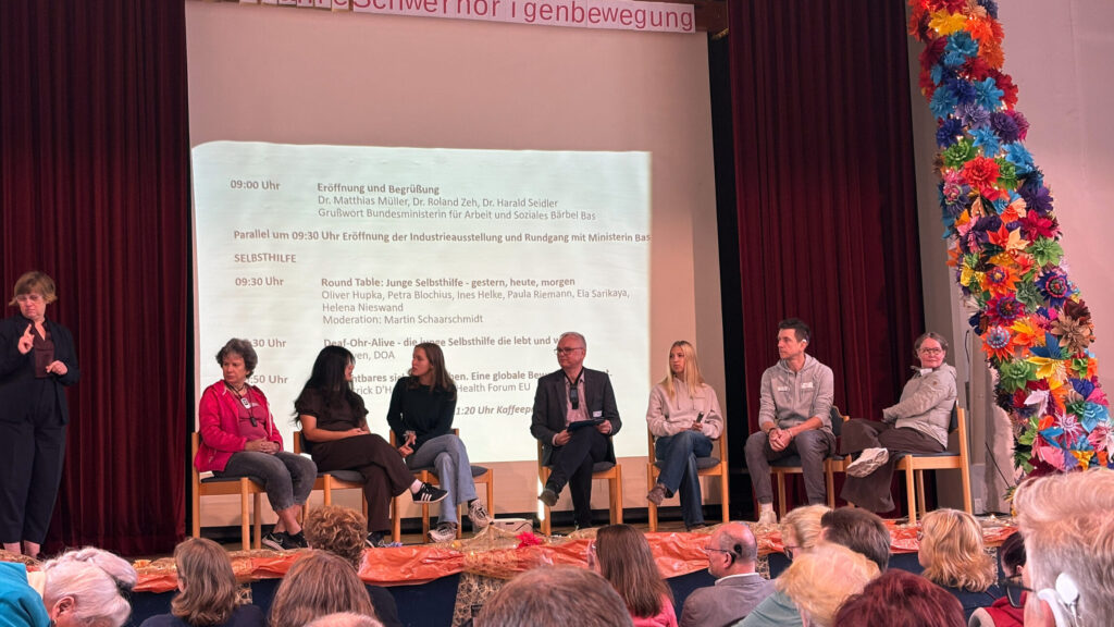 Sieben Personen sitzen auf einer Bühne vor einem Publikum. Im Hintergrund hängt ein weißes Tuch mit einer Projektion, auf der ein Veranstaltungsprogramm mit Uhrzeiten und Namen zu sehen ist. Links steht eine Frau mit Mikrofon, daneben weitere Personen in unterschiedlichen Sitzhaltungen. Rechts ist eine bunte, mit Papierblumen geschmückte Säule zu erkennen. Die Bühne ist von einem roten Vorhang eingerahmt.