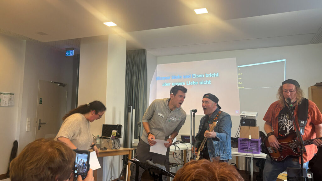 Vier Personen musizieren gemeinsam in einem Raum. Zwei Männer stehen in der Mitte vor Mikrofonen, einer hält ein Blatt Papier, der andere spielt Gitarre. Links steht eine Frau, die sich leicht nach vorne beugt, rechts ein Mann mit Bassgitarre. Im Hintergrund ist eine große Leinwand mit projiziertem Text zu sehen. Verschiedene technische Geräte und Kabel sind auf einem Tisch platziert. Einige Personen im Vordergrund sind angeschnitten sichtbar.