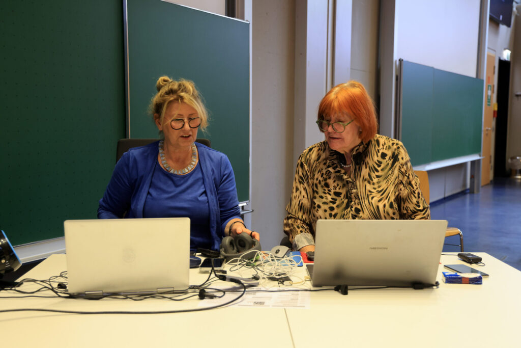 Zwei Frauen sitzen nebeneinander an einem Tisch, der mit Laptops, Kabeln und technischen Geräten belegt ist. Beide tragen Brillen; die Frau links hat blondes, hochgestecktes Haar und ein blaues Oberteil, die Frau rechts hat schulterlanges, rötliches Haar und eine gemusterte Bluse. Im Hintergrund stehen große grüne Tafeln und helle Wände.