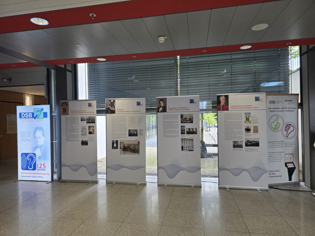Fünf große, aufrecht stehende Ausstellungstafeln mit weißen Hintergründen und blauen Schallwellenmotiven sind nebeneinander angeordnet. Jede Tafel zeigt Fotos, Textblöcke und einzelne grafische Elemente wie Piktogramme. Links steht ein weiteres, kleineres Display mit einem blauen Logo und einem Porträtfoto. Die Ausstellung befindet sich in einem modernen Raum mit großen Fenstern und grauen Jalousien.
