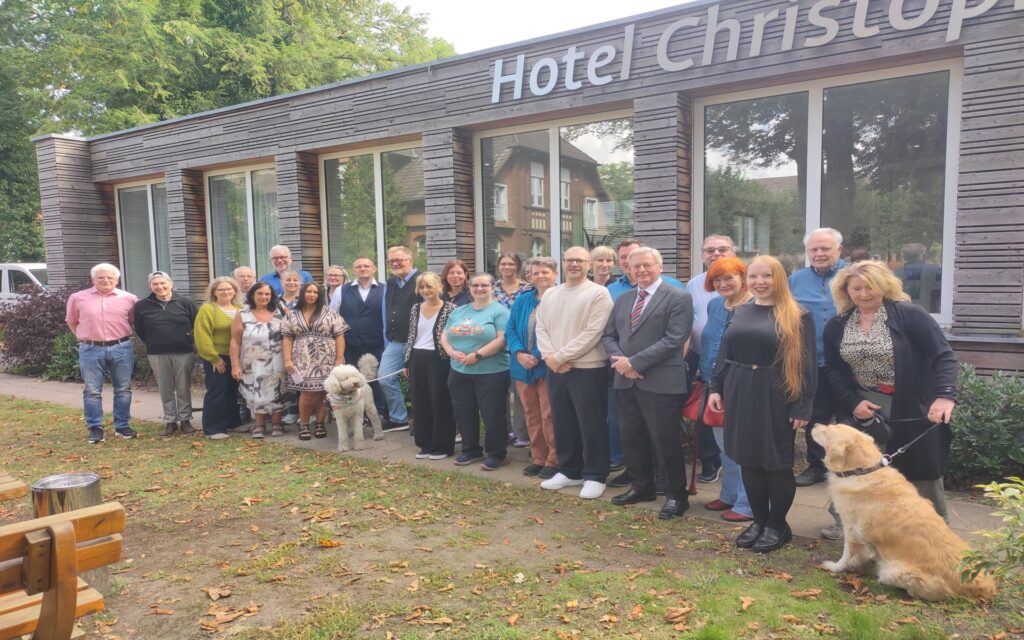Eine Gruppe von Menschen steht in einer Reihe vor einem Gebäude mit großen Fenstern. Über dem Eingang ist der Schriftzug 'Hotel Christop...' zu sehen. Einige Personen tragen formelle Kleidung, andere sind leger gekleidet. Zwei große Hunde stehen neben der Gruppe. Im Hintergrund sind Bäume und ein weiteres Gebäude sichtbar. Die Szene wirkt freundlich und entspannt.