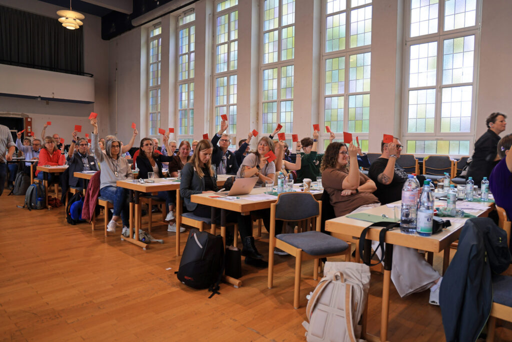 In einem großen Saal sitzen zahlreiche Menschen an Tischen, die mit Unterlagen, Getränkeflaschen und Laptops bestückt sind. Viele Personen halten rote Karten hoch. Die Fenster im Hintergrund sind groß und lassen viel Tageslicht herein. Die Atmosphäre wirkt konzentriert und gemeinschaftlich.