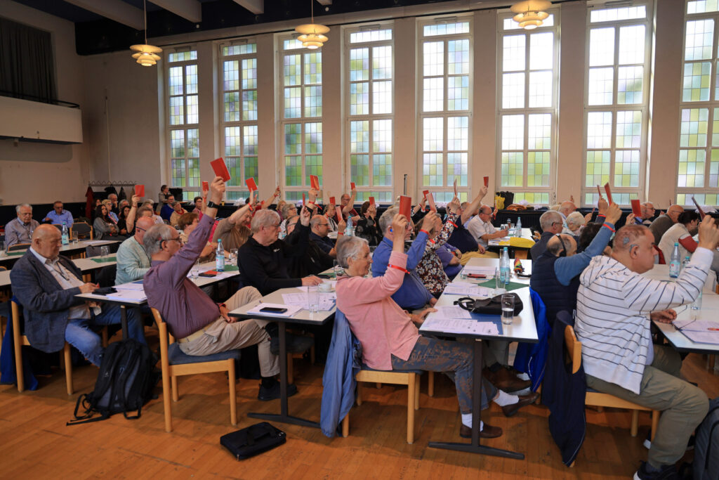 In einem großen, hellen Saal sitzen zahlreiche Menschen an langen Tischen. Viele heben rote Karten in die Höhe. Die Fensterfront im Hintergrund lässt Tageslicht herein. Auf den Tischen liegen Papiere und Schreibutensilien. Die Atmosphäre wirkt konzentriert und ruhig.