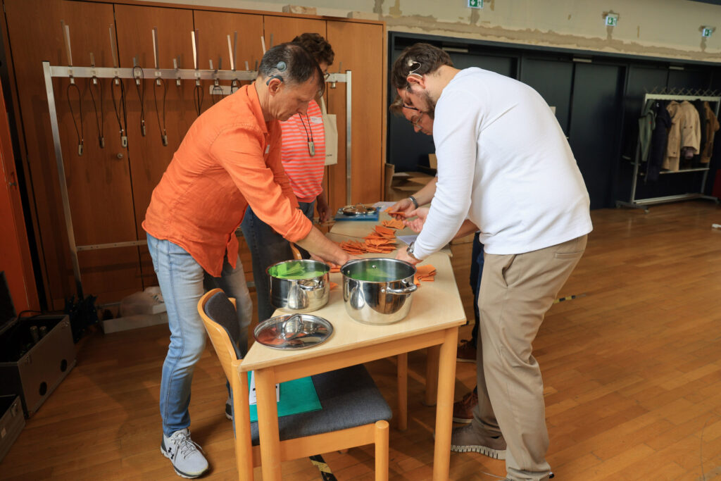 Mehrere Personen stehen um einen Holztisch in einem Raum mit Holzfußboden und hellen Wänden. Auf dem Tisch liegen orangefarbene Papierstapel und zwei große Edelstahlschüsseln. Die Personen sortieren und zählen die Papiere. Im Hintergrund sind eine Garderobe mit leeren Kleiderbügeln und einige Jacken sichtbar.