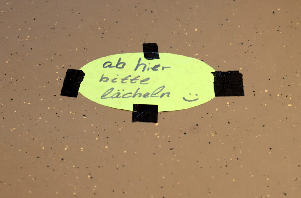 Ein ovales, neongelbes Schild liegt auf einem hellbraunen Boden und ist mit vier schwarzen Klebestreifen befestigt. In handschriftlicher Schrift steht darauf: 'ab hier bitte lächeln' und daneben ist ein kleines Smiley-Gesicht gezeichnet.