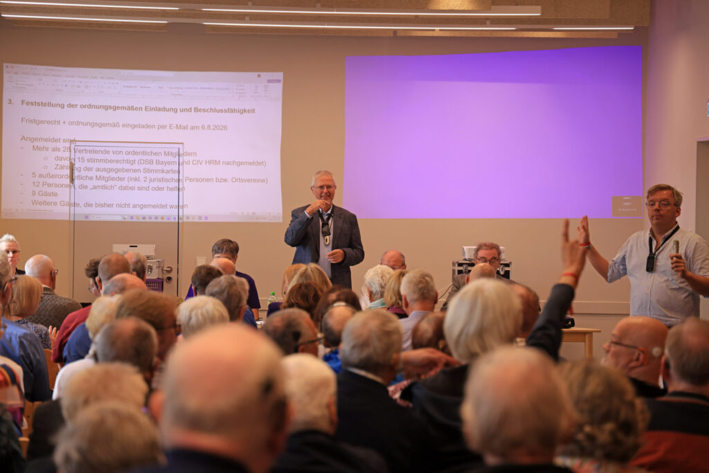 Ein Konferenzraum mit zahlreichen sitzenden Personen, die nach vorne blicken. Zwei Männer stehen vorne, einer spricht in ein Mikrofon, der andere hebt die Hand. Im Hintergrund ist eine große Leinwand mit einer Präsentation zu sehen, auf der Text in blauer Schrift projiziert wird. Die Atmosphäre wirkt konzentriert und aufmerksam.