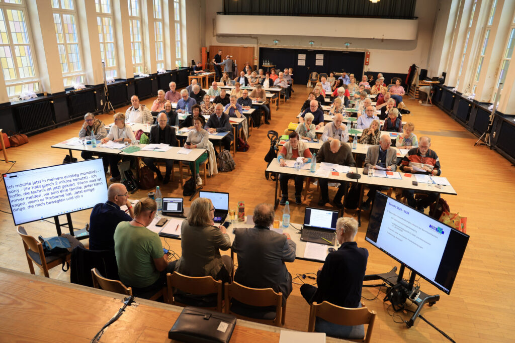 Blick in einen großen Saal mit hohen Fenstern und hellem Holzboden. Mehrere Reihen von Tischen sind aufgestellt, an denen zahlreiche Menschen sitzen und auf Laptops, Unterlagen oder Leinwände schauen. Im Vordergrund sitzt eine Gruppe an einem Tisch mit mehreren Monitoren und Mikrofonen. Zwei große Bildschirme zeigen Text und Präsentationen. Die Atmosphäre erinnert an eine Konferenz oder Versammlung.