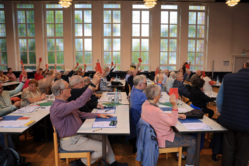 In einem großen Saal sitzen zahlreiche Menschen an langen Tischen. Viele von ihnen halten rote Karten in die Luft. Die Fenster im Hintergrund sind groß und lassen Tageslicht herein. Auf den Tischen liegen Papiere und Schreibutensilien. Die Atmosphäre wirkt konzentriert und gemeinschaftlich.