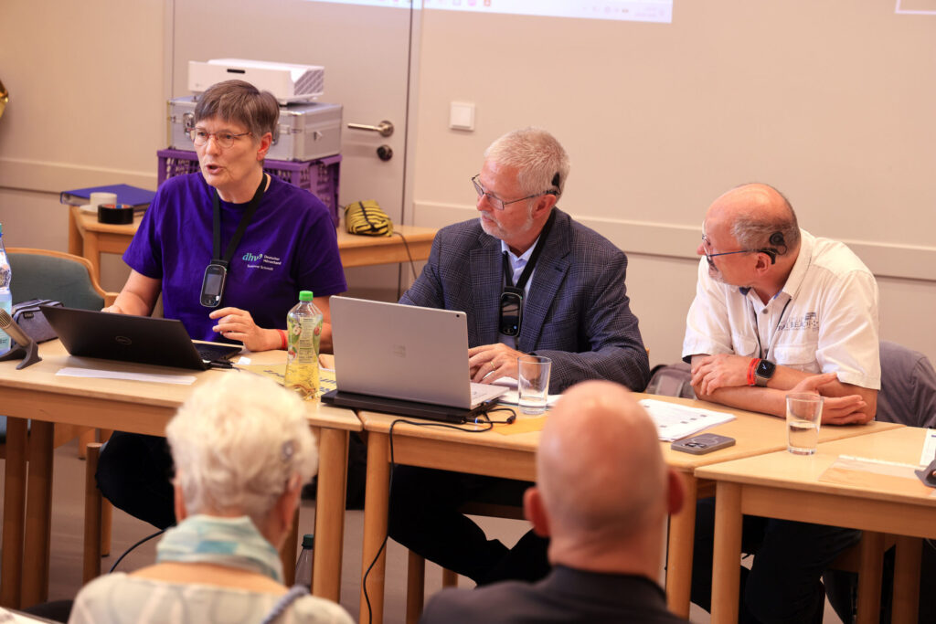 Drei Personen sitzen nebeneinander an einem langen Holztisch in einem Konferenzraum. Die Person links trägt ein lila T-Shirt und spricht, während sie auf einen Laptop zeigt. Die beiden anderen Personen tragen Hemden und Sakko, hören aufmerksam zu und blicken in ihre Richtung. Auf dem Tisch stehen Laptops, Mikrofone, Wasserflaschen und Gläser. Im Vordergrund sind unscharf mehrere Zuhörende zu sehen. Im Hintergrund befinden sich technische Geräte und eine helle Wand.