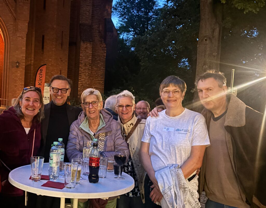 Sechs Personen stehen an einem runden Stehtisch im Freien vor einer Kirche, auf dem Tisch stehen mehrere Getränke in Gläsern und Flaschen.