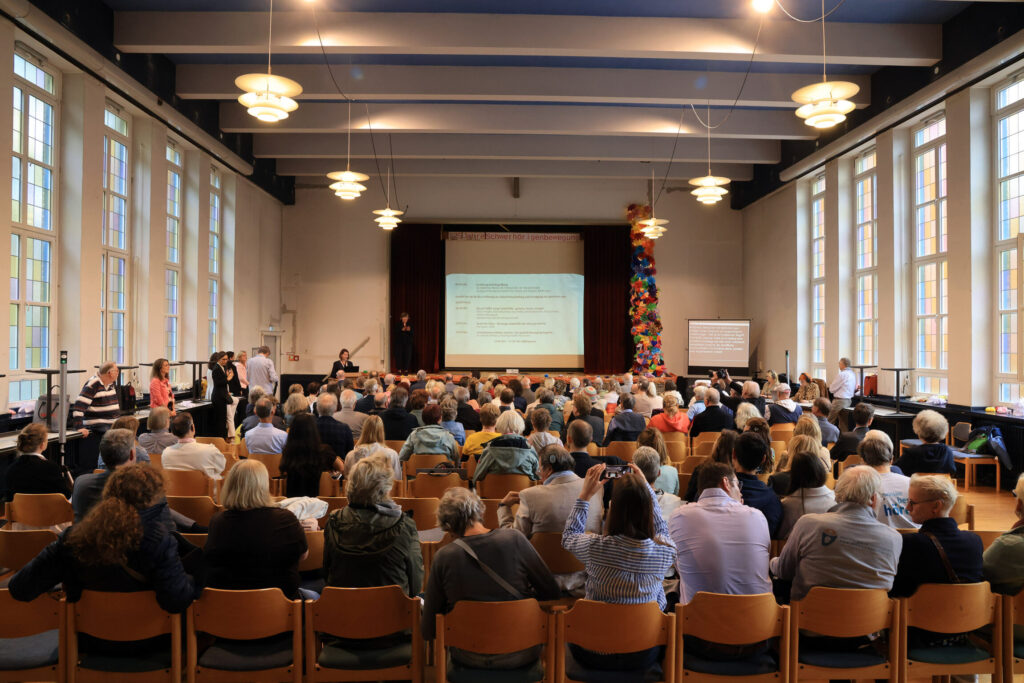 Großer Saal mit Publikum auf Stühlen. Vorne spricht am Sprecherpult eine weibliche Person vor einer Leinwand mit Präsentation und Flipchart, mehrere Deckenlampen und große Fenster an der linken Seite.