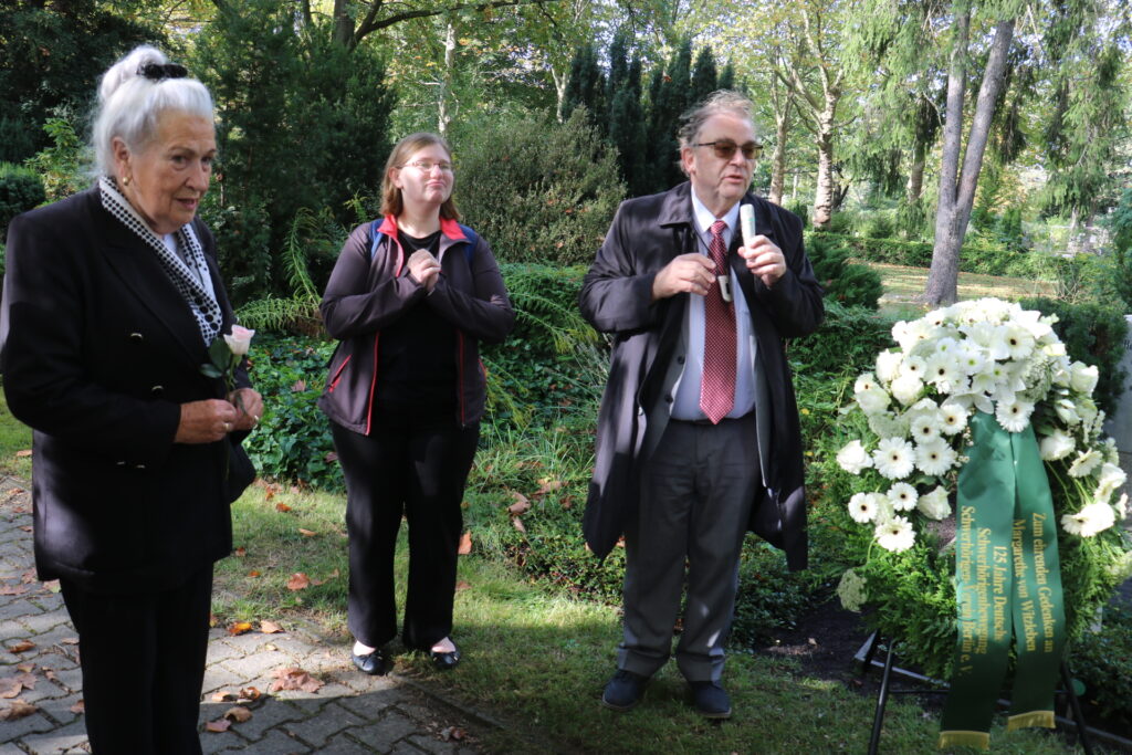 Drei Personen stehen in einem grünen Garten oder Park. Eine ältere Frau hält eine weiße Rose in der Hand und trägt einen dunklen Mantel mit einem gepunkteten Schal. Eine weitere Person steht mit gefalteten Händen und trägt eine dunkle Jacke mit roten Akzenten. Ein Mann im Anzug und Mantel spricht in ein Mikrofon. Rechts im Bild befindet sich ein großer Kranz aus weißen Blumen mit grünen Schleifen, auf denen goldene Schrift zu sehen ist. Im Hintergrund sind dichte Sträucher und Bäume.