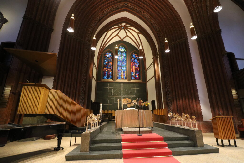 Innenraum einer Kirche mit Altar, rotem Teppich, Orgel und bunten Glasfenstern im Hintergrund.