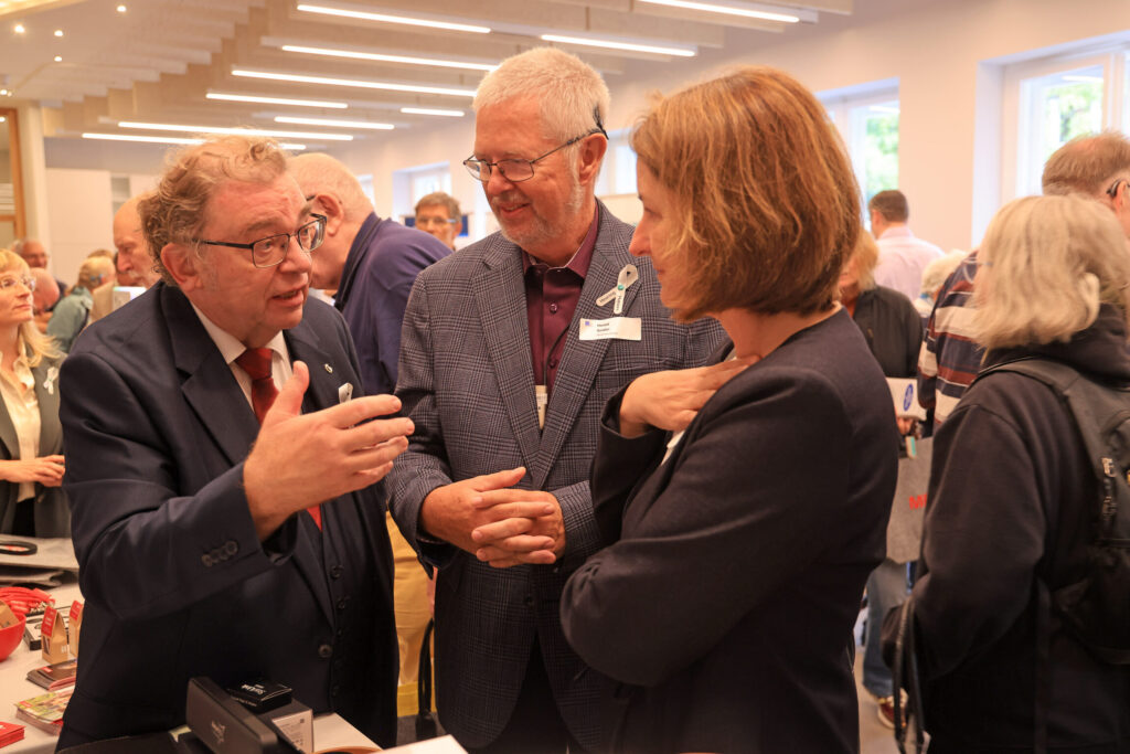 Drei Personen in einem Gespräch in einem hellen Raum mit weiteren Menschen im Hintergrund.