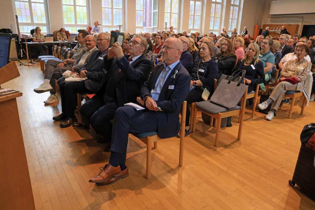 Mehrere Personen sitzen in einem hellen Raum auf Holzstühlen und hören aufmerksam zu.