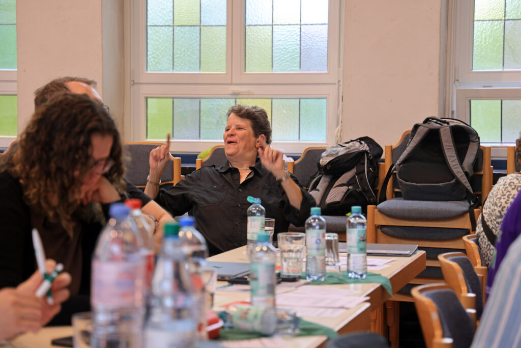Eine Person mit kurzen dunklen Haaren sitzt an einem Tisch in einem hellen Raum und hebt beide Hände mit ausgestreckten Zeigefingern. Auf dem Tisch stehen mehrere Wasserflaschen, Gläser und Schreibutensilien. Im Hintergrund sind Stühle und große Fenster mit milchigem Glas zu sehen. Weitere Personen sitzen am Tisch und sind teilweise angeschnitten.