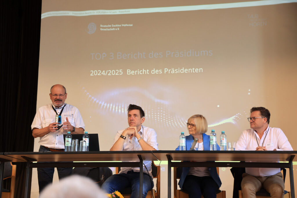 Vier Personen sitzen an einem langen Tisch auf einer Bühne. Links steht ein Mann und spricht, während die anderen drei aufmerksam zuhören. Alle tragen helle Hemden und Namensschilder. Auf dem Tisch stehen mehrere Wasserflaschen und Gläser. Im Hintergrund ist eine große Leinwand mit einer Präsentation zu sehen. Die Leinwand zeigt den Titel 'TOP 3 Bericht des Präsidiums', darunter die Jahreszahl '2024/2025' und den Text 'Bericht des Präsidenten'.