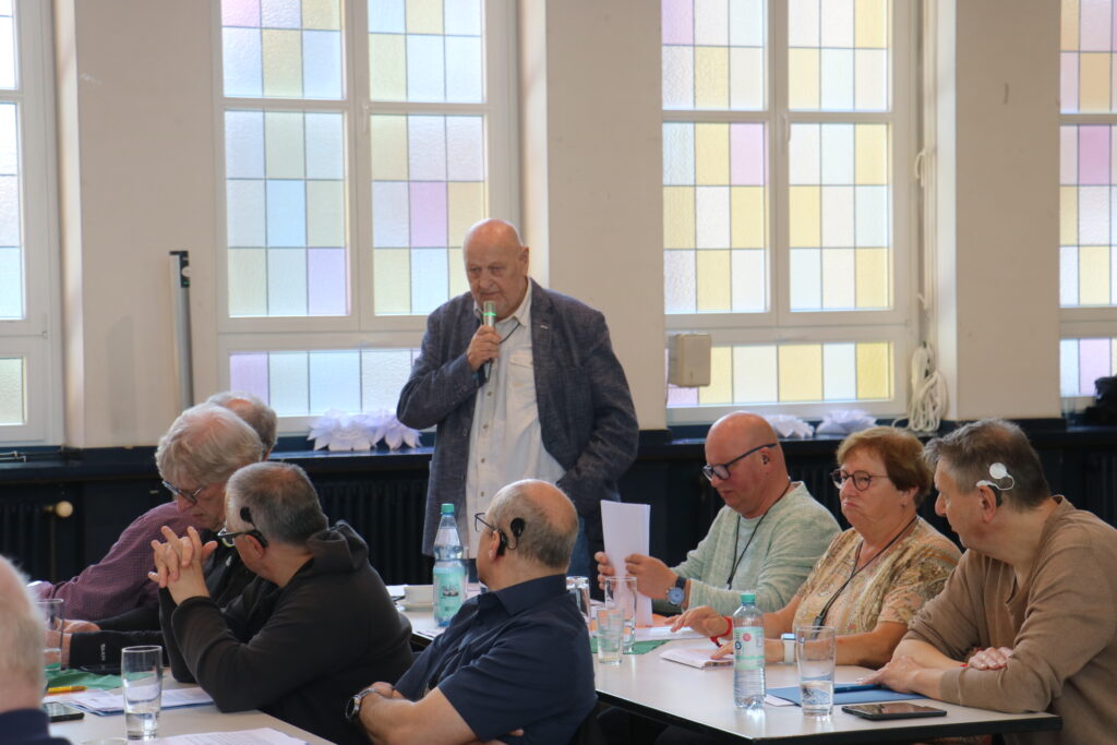 Ein älterer Mann steht in einem hellen Raum vor einer Gruppe sitzender Menschen und spricht in ein Mikrofon. Die Personen sitzen an einem langen Tisch, auf dem Wasserflaschen, Gläser und Papier liegen. Im Hintergrund sind große Fenster mit pastellfarbenen Glasflächen zu sehen.