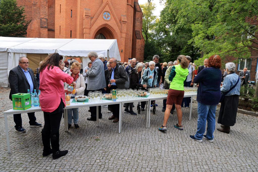 Vor einem Zelt und einer Backsteinkirche stehen zahlreiche Menschen um einen langen Tisch, der mit Gläsern und Getränkeflaschen gedeckt ist. Einige Personen schenken Getränke ein oder unterhalten sich. Die Szene spielt sich im Freien auf einem gepflasterten Platz ab, im Hintergrund sind Bäume und Gebäudedetails sichtbar. Die Kleidung der Anwesenden ist unterschiedlich, von leger bis formell.