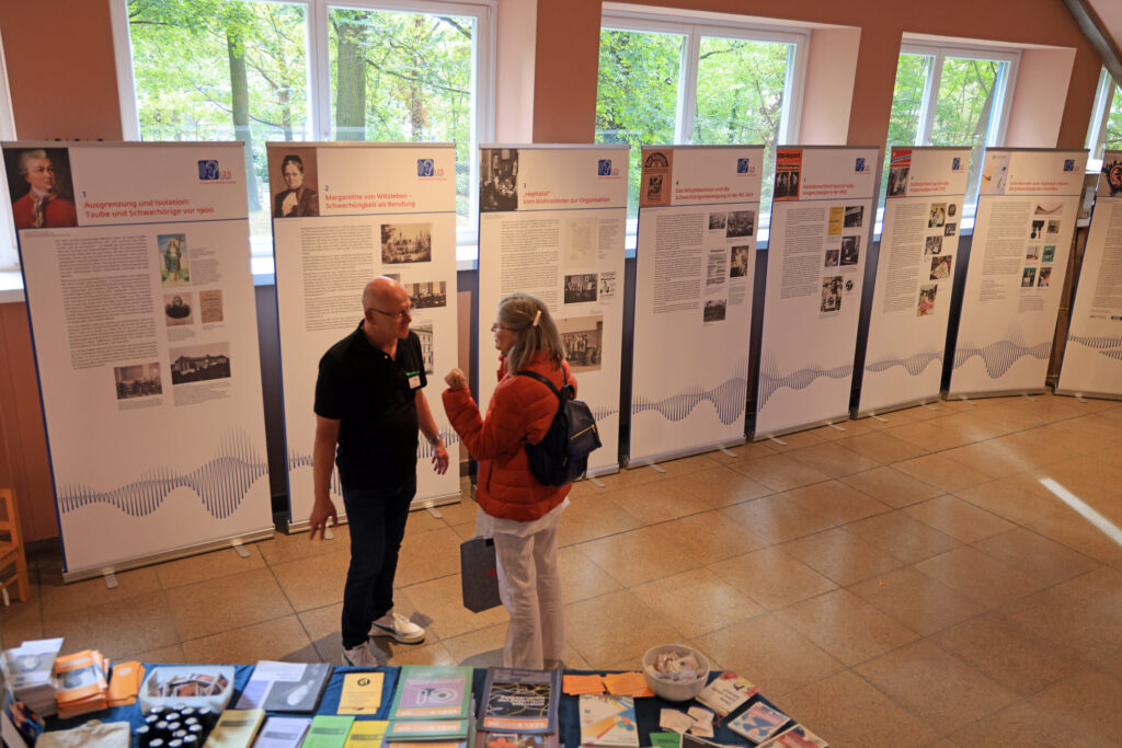 Zwei Personen stehen in einem Ausstellungsraum mit großen Fenstern und unterhalten sich. Im Hintergrund sind mehrere aufgestellte Schautafeln mit Texten, Fotos und historischen Abbildungen zu sehen. Im Vordergrund liegt ein Tisch mit zahlreichen ausgelegten Broschüren, Büchern und Informationsmaterialien in unterschiedlichen Farben. Die Schautafeln sind gleichmäßig angeordnet und zeigen Porträts, Dokumente und erklärende Texte.
