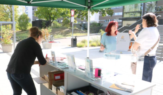 Drei Frauen an einem Infostand unter grünem Pavillon, eine Frau steht hinter dem Tisch, zwei Frauen davor im Gespräch.