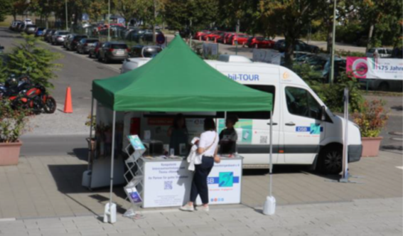 Zwei Personen stehen an einem Informationsstand mit grünem Pavillon vor einem weißen Transporter auf einem Bürgersteig.