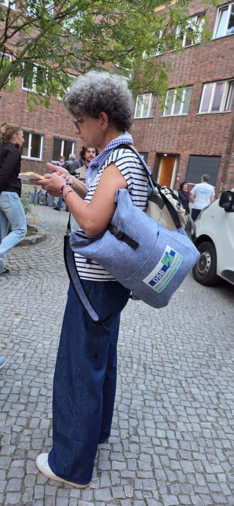 Eine Person mit grauen, lockigen Haaren steht auf einem gepflasterten Gehweg vor einem Backsteingebäude. Sie trägt eine Brille, ein gestreiftes T-Shirt und weite, blaue Hosen. Über der Schulter hängt ein großer, hellblauer Rucksack mit einem rechteckigen Aufnäher, auf dem ein Logo und Schriftzüge zu sehen sind. Die Person hält ein Smartphone in der Hand und blickt darauf. Im Hintergrund sind weitere Menschen, ein weißer Transporter und ein Baum mit grünen Blättern zu erkennen.
